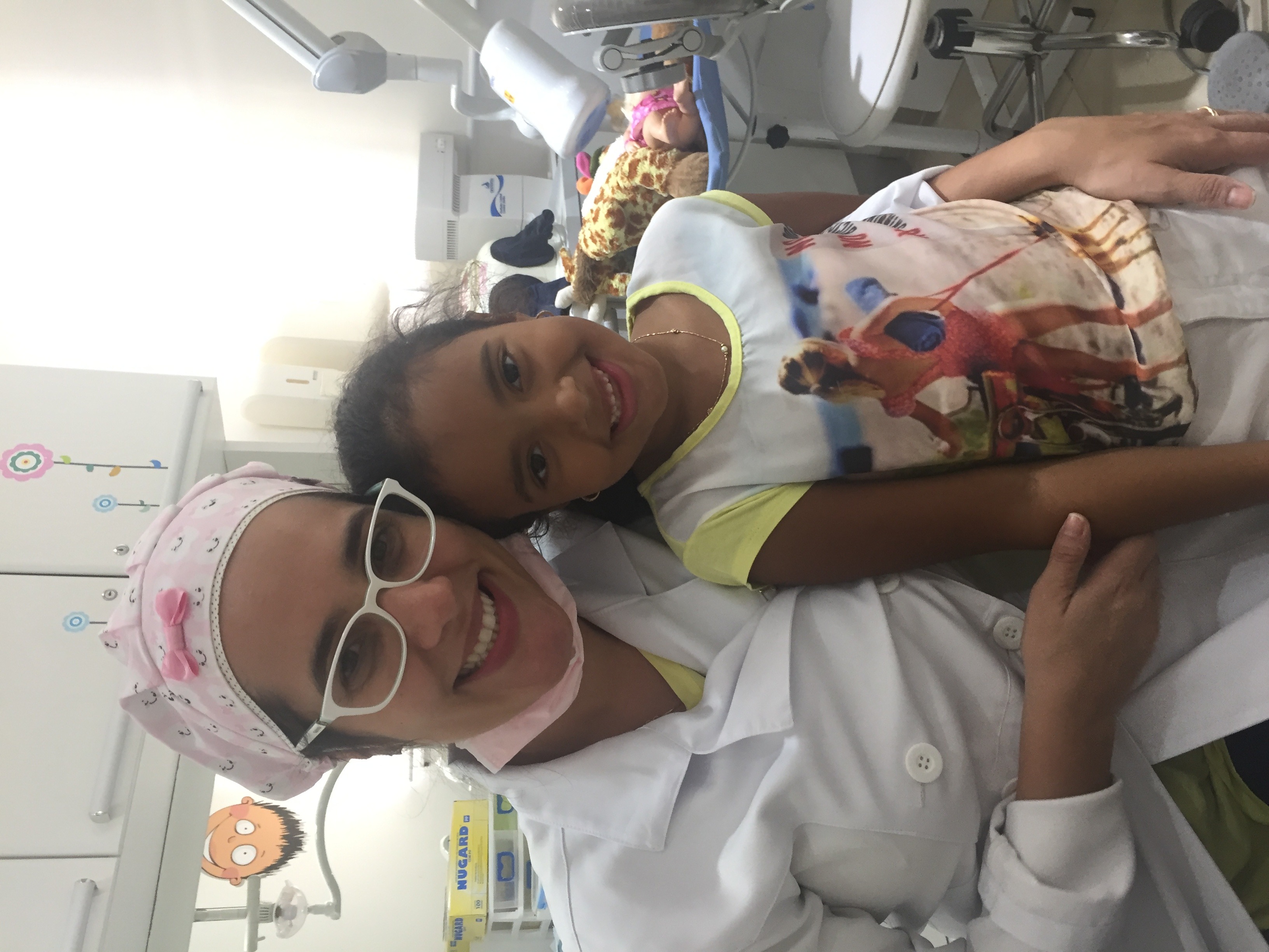 Dra. Anne de jaleco branco abraçada a uma paciente, as duas sorrindo no consultório.