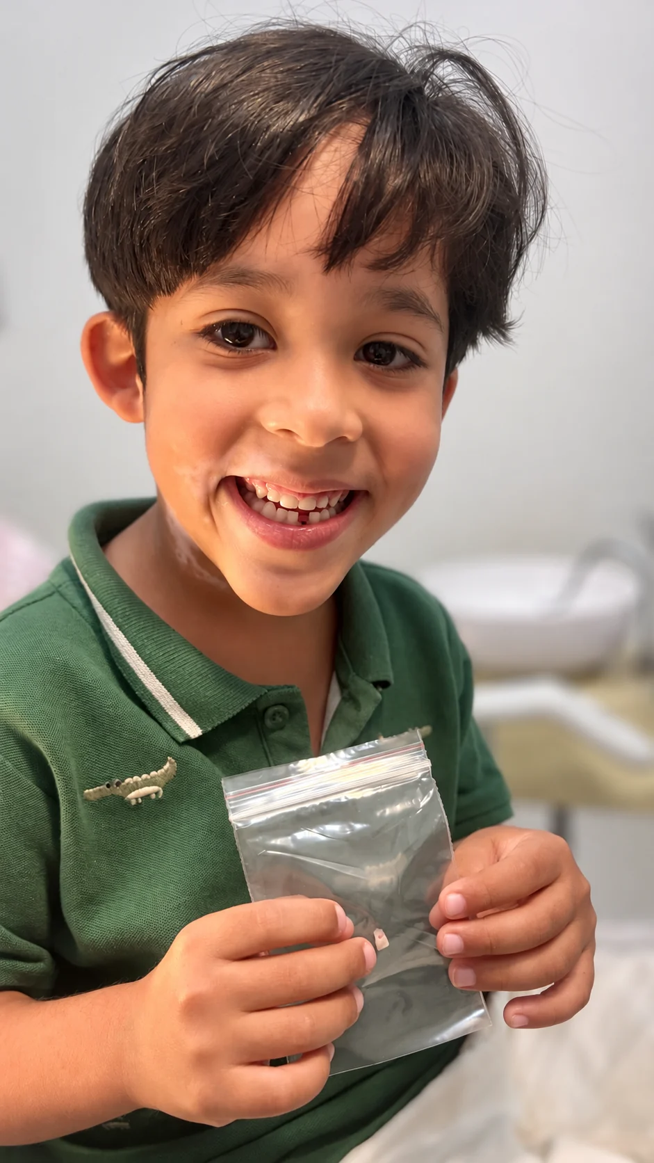 Menino de camisa verde sorrindo e segurando um saquinho no consultório.