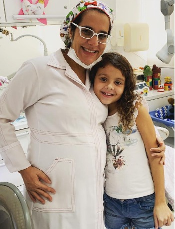 Dra. Anne de jaleco branco abraçada a uma menina de cabelo cacheado, as duas sorrindo.