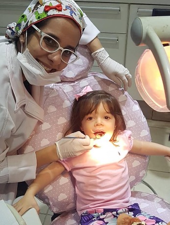 Menina com laço rosa no cabelo na cadeira do consultório, de boca aberta para o exame, com a Dra. Anne ao fundo.