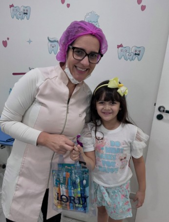 Dra. Anne de touca lilás ao lado de uma menina com laço amarelo, que segura um kit de escovas de dente.