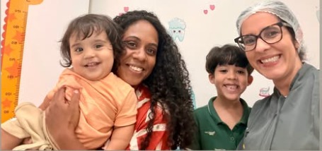 Dra. Anne com uma mãe, o filho bebê no colo e o irmão mais velho, todos sorrindo no consultório.
