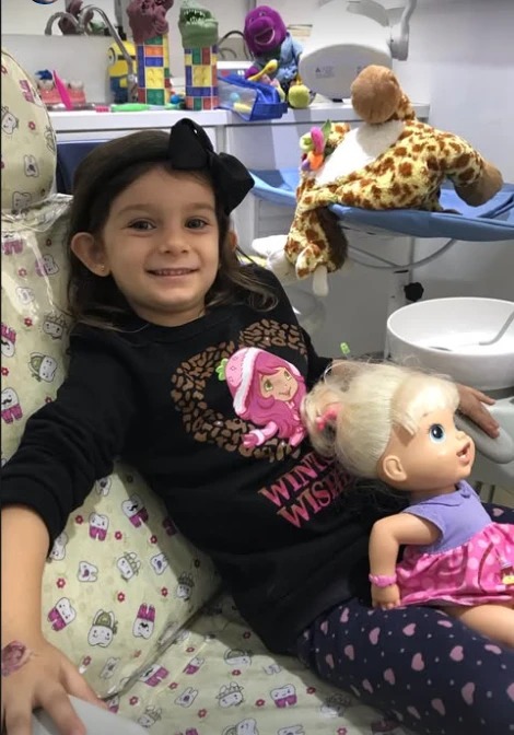 Menina com laço preto no cabelo sorrindo na cadeira do consultório, abraçada à sua boneca.