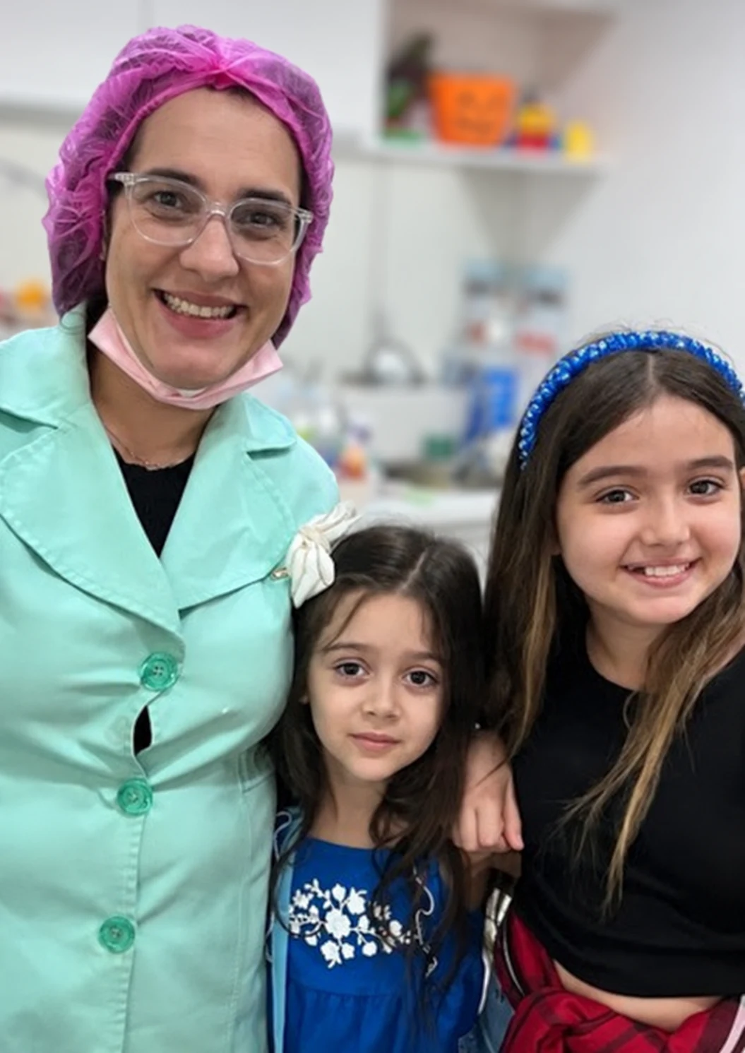 Dra. Anne de jaleco verde ao lado de duas primas sorrindo no consultório.
